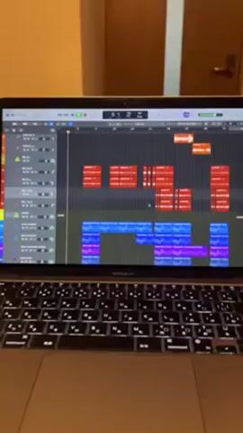 The video shows a close-up of a MacBook Air laptop with a music production software interface displayed on the screen. The software interface shows a multi-track audio project with several colored blocks representing different audio segments. Red blocks are prominent at the top, while blue and purple blocks are arranged beneath. The laptop's keyboard is visible in the foreground, and the device rests on a wooden surface. In the background, part of a beige-colored wall and a closed door are visible. The focus is primarily on the laptop screen and the audio production software.