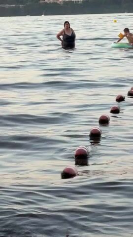 A woman in a black swimsuit stands in the water, facing away from the camera with her hands on her hips. To her right, a child wearing arm floaties sits on an inflatable raft in the water. A string of red and white buoys stretches across the water from the foreground to the background, marking a boundary. The water is rippled, and a tree line is visible in the distance.