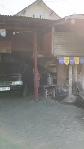 The video shows a person working on a black vehicle parked in what appears to be an open-air garage or workshop. The vehicle is a Suzuki Carry with license plate number L 9592 GE. The person, partially obscured, is engaged in some activity related to the vehicle's engine or undercarriage. In the background, there is a large blue water jug on a stool, and various other items, including what looks like shelving and building materials. The ground is paved with bricks. Sunlight is visible, suggesting it is daytime.