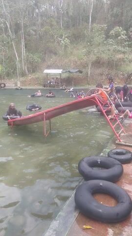 The video shows a shallow pool with murky water and a red slide. Several people are in the pool, some on inflatable tubes. The slide has a red ladder leading up to it. In the background, there is lush green foliage and trees. There are also some black inflatable rings lying on the edge of the pool. The overall impression is of a casual, outdoor recreational area.