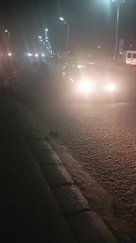 A shaky, first-person perspective video shows a nighttime street scene. Street lights illuminate the wet asphalt road, where several cars are traveling. The bright headlights of oncoming vehicles are visible through the haze. A dark curb runs along the left side of the road, with dark shadows and indiscernible objects nearby. The sound of a car horn is audible.
