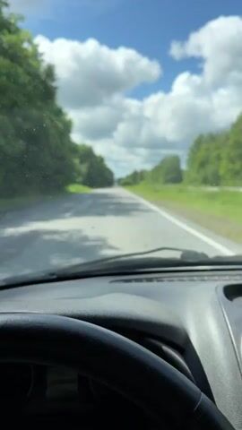 The video begins with a first-person perspective shot from inside a car, looking out the windshield. The road ahead is paved and appears to be a two-lane highway or country road, flanked by lush green trees on both sides. The sky is a bright blue with fluffy white clouds. The car is moving, indicated by the motion blur of the trees and the road.

The perspective then shifts to a close-up of the car's dashboard. A speedometer is visible, with the needle indicating a speed of approximately 120 km/h. To the left of the speedometer is a digital display showing the time as 14:25, the odometer reading as 167069 km, and the fuel consumption as 8.5 L/100km. A fuel gauge is also present, showing the tank is nearly full. The dashboard appears to be made of dark plastic.

The video concludes by returning to the first-person view of the road ahead, with the car continuing its journey through the scenic, tree-lined landscape under a bright, cloudy sky.