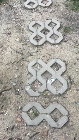 This media file shows a close-up, slightly overhead view of a garden path constructed from concrete pavers. The pavers are of a unique interlocking, octagonal design, arranged in a staggered pattern to form a winding walkway. The path is bordered by patchy grass and a dirt/gravel ground scattered with small twigs and leaves. The overall impression is of a somewhat rustic or natural outdoor setting, possibly a garden or a less-maintained area of a park.

The pavers are the primary focus. They are a light gray concrete, with a distinctive shape that resembles two interlocking figure-eights or infinity symbols. The pattern repeats down the length of the path, with a few of the pavers showing signs of wear or minor damage, such as chips or smudges. The arrangement creates a sense of movement and direction, leading the viewer's eye further into the scene.

The ground surrounding the pavers is a mix of soil, small stones, and debris, indicating an outdoor environment that may not be regularly manicured. Patches of green grass are visible on either side of the path, suggesting it is an area where vegetation is present. The lighting appears to be natural daylight, with no harsh shadows, suggesting it could be a cloudy day or perhaps late afternoon. The scene is devoid of people, animals, or any other distinguishing features, focusing solely on the path and its immediate surroundings. The location context of Iași, Romania, suggests this is a European setting. There is no visible text in the media file.