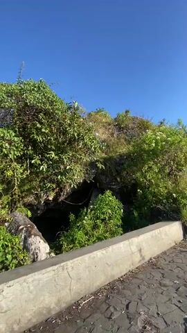 The video pans across a scenic mountainous landscape under a clear blue sky with the sun shining brightly. In the foreground, a paved path runs alongside a concrete barrier, bordered by lush green vegetation. As the camera moves, it reveals a breathtaking panoramic view of a valley filled with houses and agricultural fields. The sun casts a warm glow over the rolling hills, creating a serene and picturesque atmosphere. The time of day appears to be morning or late afternoon, given the angle of the sun and the soft light. There are no people or specific objects prominently featured, emphasizing the natural beauty of the location. The setting is clearly rural, with the village nestled amidst the natural contours of the land. The overall mood is peaceful and awe-inspiring, showcasing the grandeur of the Indonesian countryside. There is no visible text in the media file.