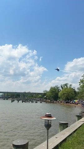 The video depicts a clear, blue sky with scattered cumulus clouds, against which a dark-colored jet aircraft performs an aerial maneuver, leaving a white contrail. The jet is seen from multiple angles as it flies over a body of water. In the background, a bridge spans the water, and a shoreline is visible, populated with people, piers, docks, boats, and lampposts. In the foreground, a wooden dock is visible with lamp posts and bollards. The scene is bright, indicating daytime, and the overall impression is one of a public event involving an air show over a waterway.