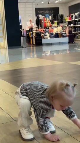 The video captures a scene inside an airport, likely in Перепечино, Russia, with a focus on a young child. The child, with blonde hair styled in two pigtails, is wearing a striped long-sleeved shirt and light-colored corduroy pants. They are looking off to the side with a curious expression. The background reveals a Samsonite store filled with various luggage items and accessories. A woman can be seen standing behind the store counter, interacting with a customer or attending to her work. The floor is tiled with a wood-like pattern, reflecting the overhead lighting. Another person is walking past in the background, also appearing to be a traveler. The overall atmosphere suggests a typical day at an airport, with people moving about and shops open for business. The time of day is not specified but the indoor lighting suggests it could be daytime.