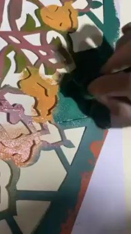 A pair of hands dabs at a piece of stencil with a green sponge. The stencil features a repeating geometric pattern with a layer of colors applied to some sections of the stencil. Below the stencil is a piece of white paper. The hands move the sponge in a dabbing motion over the stencil, applying green paint to the areas exposed by the stencil’s cutouts. The stencil has previously been used, and shows signs of wear and paint residue.