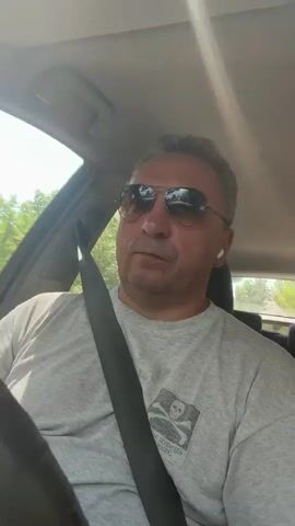 A middle-aged man wearing sunglasses and a grey t-shirt with a skull and crossbones logo is driving a car. He has a seatbelt on and is wearing white wireless earbuds. The sun is shining brightly, creating glare on the windshield. Trees and foliage are visible outside the car windows, suggesting an outdoor environment. The man is looking forward, presumably at the road, and appears to be speaking or engaged in some activity inside the car.