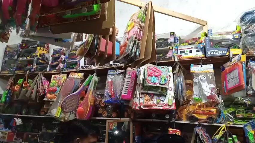 The video shows the interior of a small shop selling a variety of toys and novelty items. The shelves are packed with merchandise, primarily in plastic packaging, including toy cars, toy guns, toy keyboards, and small stuffed animals. Some items hang from the ceiling or are displayed on stands. Two individuals are partially visible in the foreground, appearing to be customers or staff. The lighting is somewhat dim and appears to be artificial, coming from overhead fixtures. The overall impression is of a cluttered but well-stocked retail environment.