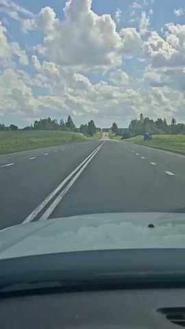 The video shows a view from the dashboard of a car driving on a paved road. The road is straight with two white lines marking the lanes. On the left side of the road is a field of crops, possibly corn. On the right side are trees and a blue structure. The sky is blue with fluffy white clouds. The car's hood is visible in the foreground.
