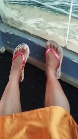The media file shows a first-person perspective of someone's legs and feet wearing white flip-flops with pink straps. The person is wearing an orange skirt or dress. The scene appears to be outdoors, with a dark asphalt-like ground visible. In the first few seconds, there's a glimpse of a blue and white structure with a window, possibly indicating a doorway or a building. The rest of the footage shows the person's legs and feet against the dark ground, with a blurred background that suggests movement. The lighting indicates it's daytime, and there are no specific weather cues. The overall impression is of someone walking or moving, possibly in a casual setting given the footwear. There is no visible text in the media file. The context of Uselu, Nigeria, suggests an urban or semi-urban environment within that region.