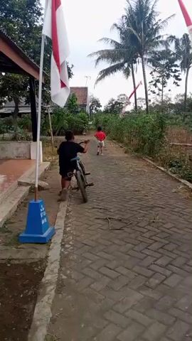 A young boy in a black shirt and shorts pushes a bicycle along a paved pathway lined with red and white Indonesian flags. He is moving away from the camera, towards a cluster of trees and buildings. Another child, wearing a red shirt and shorts, is further up the path, also moving away from the camera and appears to be riding a bicycle. The environment is outdoors, with a cloudy sky, palm trees, and dense vegetation.