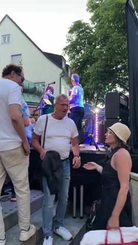 A group of people are gathered in front of a stage where a band is playing. The stage is lit with purple and red lights. A man in a white t-shirt and jeans is standing on the left side of the stage, playing a guitar. He is wearing glasses and has short, gray hair. Another man in a white t-shirt is standing next to him, and a woman in a dark dress and hat is standing on the right side of the stage. In the foreground, a man and woman are holding hands and walking down some steps. The man is wearing a white t-shirt and jeans, and the woman is wearing a black dress and a hat. There are trees and buildings in the background.