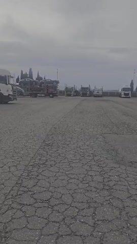 The video shows a cracked asphalt lot with several car carrier trucks parked in the background. On the left, a red car carrier truck is loaded with two cars. To the right of it, three more car carrier trucks are parked in a line. The sky is overcast.