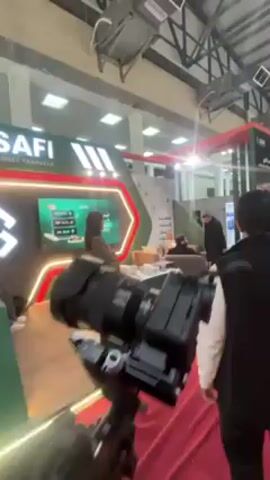 The video shows the interior of a convention hall, with a prominent booth for "SAFI Money Transfer." The booth is decorated in green and red and features a reception desk with the SAFI logo. Behind the desk is a large screen displaying information. A woman stands near the desk, and several men are seated in chairs in a lounge area within the booth, appearing to be in conversation. The camera is mounted on a gimbal and moves smoothly, showing the booth from different angles. The floor is covered with a red carpet. The hall is well-lit with overhead lighting. Other booths and people can be seen in the background, but they are less focused.