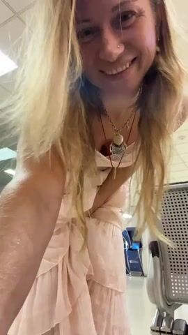The video shows a woman with long blonde hair wearing a light pink lace bralette and a long, layered pink tulle skirt. She is standing in what appears to be an indoor public space with a tiled ceiling and fluorescent lighting. To her right is a grey metal chair with a perforated seat. The woman is in the process of spinning, causing her skirt to fan out around her. She is smiling and looking towards the camera. In the background, there are glass partitions and some blue equipment.