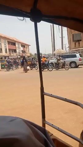 From inside a yellow tricycle taxi, the view shows a busy intersection in a city in Africa. The road surface is brown. Numerous motorcycles, several carrying passengers, move in various directions through the intersection. Several other tricycle taxis, mostly green and yellow, also navigate the intersection. A silver SUV is visible amongst the traffic. Pedestrians walk along the side of the road.  Buildings line the streets in the background, some multiple stories high and with visible shops. Power lines run overhead.  The sky is bright and clear. The camera is shaky due to the movement of the tricycle taxi.  A hand is visible resting on the taxi's metal bar in the lower left corner of the frame.