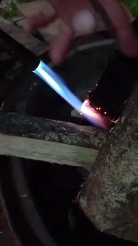 A hand holds a torch with a blue flame to a dark, rough surface, possibly wood. Small sparks fly from the point of contact. Another piece of light-colored wood is briefly brought into the frame near the torch. The scene appears to be outdoors at night, with some greenery visible in the background. The background is dark, and the primary light source is the torch flame.