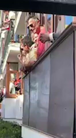 The video depicts a daytime scene on a sunny day in what appears to be an urban street or square in Vejer de la Frontera, Spain. Multi-story buildings with balconies line the area, and crowds of people are visible, both on the street level and watching from the balconies above.

Initially, several people are seen leaning over a balcony railing in the foreground, looking down at the street. People on other balconies further down the street are also watching. On the street level, a crowd of people is gathered.

Suddenly, a large, dark-colored animal, identified as a bull, emerges from the right side of the frame, near a line of green hedges or bushes. The bull quickly moves into the bushes, where it appears to lie down or roll, becoming partially obscured by the foliage. Upon the bull's appearance, the crowd on the street reacts, with some people quickly moving away from the area where the bull appeared. People on the balconies continue to observe the event. No visible text is discernable in the video.