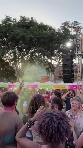 A crowd of people celebrate the Holi festival at an outdoor event with a stage, food stands, and a tall modern building in the background. The crowd is diverse, with people of different ages, ethnicities, and appearances. Many are covered in colorful powder. They dance, sing, and take pictures. On stage, a man in traditional Indian attire sings into a microphone. The atmosphere is festive.