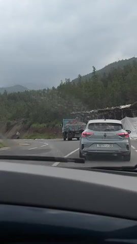 The video captures a forward-facing view from inside a moving vehicle, looking out through the windshield. The scene depicts a winding mountain road under an overcast and grey sky, suggesting cloudy or potentially rainy weather.

In the foreground, parts of the recording vehicle's dashboard are visible, including air vents. Ahead, a light grey Renault Clio car is prominent, followed by a blue dump truck. The truck is carrying a substantial load of logs or cut wood. The Renault Clio's Moroccan license plate, "78771 و 26", is clearly visible, as is the "CLIO" badge on its trunk. A "60" sign, likely indicating a speed limit, can be faintly seen on the rear of the blue truck.

The road itself appears wet in places, and a road sign indicating a right curve is briefly visible on the left. The surrounding landscape consists of hills and mountains densely covered with green trees and vegetation. On the right side of the road, some rudimentary structures or stalls covered with white tarpaulin are passed.

Throughout the video, music is audible, indicating it is playing within the recording vehicle. The overall setting is a mountainous, forested region consistent with the provided location context of Taljarft, Morocco.