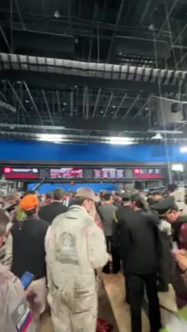 The video shows a crowd of people, most of whom appear to be covered in fake blood, suggesting a zombie-themed event. The scene takes place in a large indoor space with high ceilings, resembling a train station or airport terminal, with illuminated flight or train schedule boards visible in the background. The people are densely packed, walking around, and talking. Some are wearing costumes, including jumpsuits and business attire. The lighting is bright, with red lighting fixtures hanging from the ceiling. The general atmosphere suggests a special event or performance.