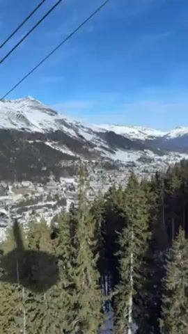 The video depicts a view from a cable car ascending towards snow-covered mountains on a clear, sunny day. The camera captures the scene through the cable car window, with the support cables visible in the frame. Below, a town nestled in a valley is partially covered in snow, surrounded by dense forests. The focus is on the picturesque mountain scenery and the aerial perspective provided by the cable car.