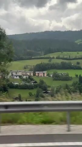 The video shows a blurry, side-view perspective from a moving vehicle. A metal guardrail runs horizontally across the lower portion of the frame, separating the road from a grassy shoulder and then a dense line of green trees. In the background, there are rolling hills covered in forests, with some glimpses of buildings in a valley. In the far distance, hazy mountains are visible under a cloudy sky. The motion blur suggests the vehicle is traveling at a moderate to high speed.