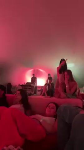 The video shows a dimly lit space with a red ambiance, where a group of people is gathered. Some are seated on sofas, appearing relaxed and attentive, while others stand behind a low platform where performers are visible. The performers include a person with a microphone and other individuals potentially operating equipment. The lighting seems to focus on the performers, creating a brighter area behind them. The setting suggests a concert or performance in an intimate, lounge-like environment.