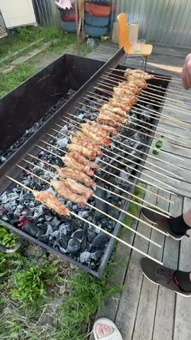 A high-angle shot captures an outdoor barbecue scene. In the foreground, a person's feet in white slides and black shoes are visible on a wooden deck. An old metal barbecue grill, black with a rustic appearance, is filled with dark coals. Numerous wooden skewers, loaded with chicken pieces, are arranged across the grill. The person's hands are turning the skewers, cooking the chicken. In the background, a small portion of a house with a corrugated metal wall and a green lawn is visible. An orange plastic chair stands next to the house, and a few plastic barrels are stacked near it.