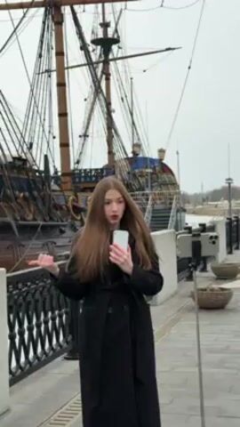 A young woman with long brown hair, wearing a black overcoat, is standing outside, appearing to be recording a video with her cell phone. She gestures towards a large, historical-looking ship behind her, mentioning that it's a Russian battleship. Another phone is attached to a tripod. The background suggests a dock or waterfront area. It's a cloudy day.