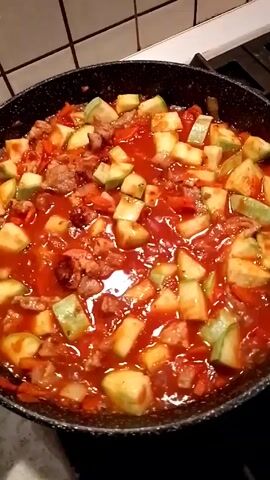 The video shows a black pan filled with a vibrant red tomato sauce, mixed with diced zucchini and small pieces of meat. The pan is on a stovetop with white-tiled walls in the background.  The food appears to be cooking or simmering.  A voice says "Mmm, how good it smells."