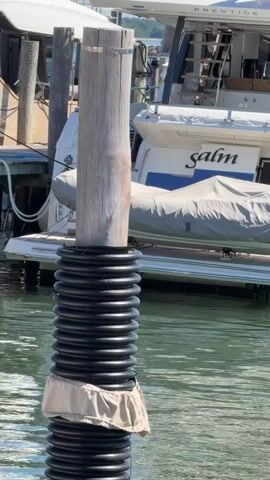 A vertical wooden post with a metal band around its top is surrounded by a ribbed black plastic pipe. The pipe is submerged in green water. A beige fabric piece is draped around the bottom of the pipe. Behind the post, a white boat with "salm" written on its side is docked. Other boats and a wooden dock are also visible in the background. The sun is shining.