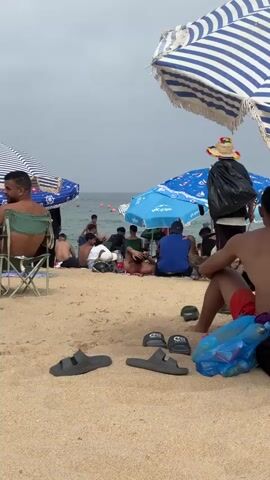 The video shows a crowded beach with people sitting under umbrellas on the sand. A person wearing a hat and carrying a large black bag walks through the scene. A goose is present among the people. Several pairs of sandals are visible on the sand in the foreground. The ocean is in the background.