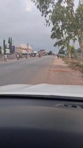 The video shows a view from inside a car looking out at a street in a town. The street is asphalt, with a shoulder of dirt on the right. There are people walking on the side of the street and buildings on the left. A yellow auto rickshaw is driving toward the car. The sky is overcast. There are trees on the right side of the road.