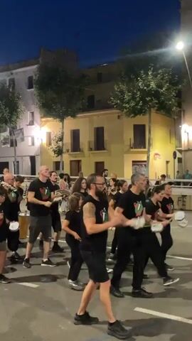 A group of people, many wearing black t-shirts with a red, white, and green emblem, are parading down a street at dusk. Some individuals are playing drums, while others are holding and striking tambourines or similar instruments. The people are in motion, some dancing or moving rhythmically. Buildings with lit windows line the street, and trees are visible. The sky is dark blue, indicating it is evening.