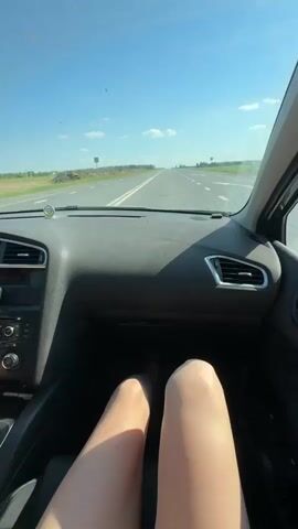 Here is a description of the video:

The video is shot from the interior of a car driving on a straight, two-lane road on a sunny day. Fields can be seen on either side of the road. The camera is positioned on the dashboard, with a view of the road ahead. Initially, the view is obscured by a person's legs, which are resting on the dashboard. A hand then enters the frame, holding up a clear plastic cup filled with an orange-yellow liquid and a black straw. The hand is wearing a white smartwatch and a bracelet. The drink is held up for a few seconds before being lowered again, with the view returning to the road.
