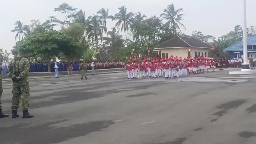 A marching band composed of young people in red and white uniforms with red hats is parading on a paved outdoor area. They are carrying instruments, including drums. Two individuals in camouflage military uniforms are standing and observing the parade. In the background, there is a line of spectators, some of whom are students in uniform. Trees, palm trees, and buildings are visible in the background. The sky is overcast.