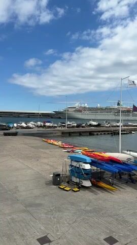 The camera films a coastal dockside location, under a partly cloudy sky. A large cruise ship looms in the background behind a harbor filled with assorted smaller boats. Closer to the camera is a paved area where a large number of colorful kayaks are stored. Several people are present, standing and walking around near the boats. A flagpole with a white flag bearing the words "PORTO DA MADEIRA"  is visible. Overall, it is a calm, sunny day at a bustling harbor facility.