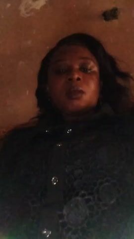 A woman is lying on her back. She is wearing a dark lace shirt with buttons. Her hair is dark. The ceiling above her is brown and textured.  The video is shaky.