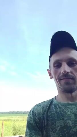 A man wearing a green camouflage t-shirt and a black baseball cap smiles directly at the camera. He is outdoors, with a bright blue sky and some trees in the background. A field of grass and what appears to be a building or greenhouse are visible in the distance. The camera appears to be held by the man himself, suggesting a selfie video.