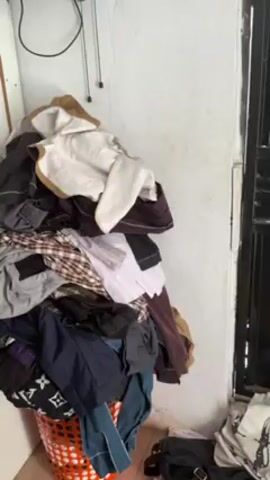 The video shows a laundry area. A basket overflowing with clothes sits next to a washing machine and a closed, barred door.  A black bag and white t-shirts lay on the floor near the door. The camera pans to an ironing board with a white sheet and an iron resting on it. A hand touches the sheet.