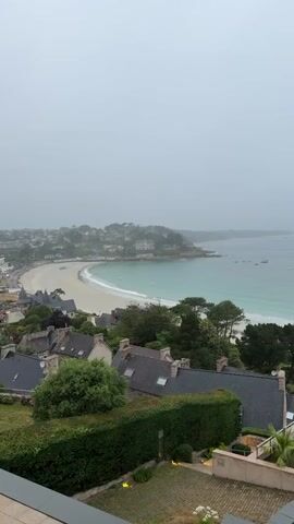 This is a static, vertical, high-angle shot of a coastal town on a cloudy day. In the foreground, there are several houses with dark-tiled roofs and lush green hedges. A small patio area with a table and chairs is visible on the right. Beyond the houses, a crescent-shaped sandy beach borders a turquoise body of water. The water is calm with a few small boats dotted on the surface. In the distance, more houses and buildings line the coastline, and the land recedes into a hazy, overcast sky. The overall atmosphere is muted and tranquil. No people are visible in the frame.