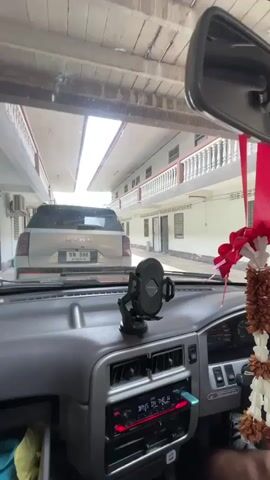 The video shows the interior of a car from the driver's perspective. The driver is following another car, a silver SUV, through a covered walkway or alleyway. The walkway is lined with apartment-like buildings on both sides. The buildings have balconies with white railings and air conditioning units visible. The sun is shining brightly at the end of the walkway. The dashboard of the car is visible in the foreground, with a phone mount and a car stereo. The other car has a license plate with Thai characters. The audio is a conversation in Thai, with one person asking if the other is going to work.