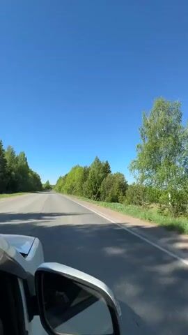 A white car drives on a grey asphalt road under a clear blue sky. The car is heading towards the horizon, where the road appears to disappear into the distance. Dense green trees line both sides of the road, forming a thick forest. There are no other vehicles visible on the road. A sign is seen beside the road. The sound of static is heard.