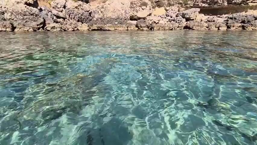 This video captures a scenic view of a clear turquoise sea from a low vantage point, likely from within the water. The water is transparent, allowing visibility of the sandy bottom and shimmering light patterns on the surface. In the background, a rocky coastline is visible, topped with sparse vegetation. Further in the distance, a beach with numerous people enjoying the sun and sea can be seen. Buildings and trees are also present along the coastline, suggesting a developed resort or tourist area. The sky is clear and blue with a few scattered clouds, indicating a bright, sunny day. The overall impression is that of a serene and inviting coastal environment.