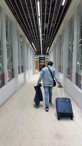 A man walks away from the camera in a long corridor with a suitcase and bag. The corridor has white walls with large windows. The ceiling is lined with horizontal wooden beams with lights in between. He is wearing jeans, a light blue jacket, and a dark shoulder bag. He walks towards a sign that says "Betraco" in the distance. The floor is light with a subtle pattern.