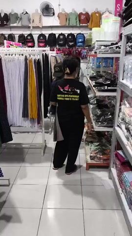 The video shows the interior of a clothing store. In the foreground, a person wearing a black t-shirt with graphics and black pants is standing, facing away from the camera, browsing a rack of clothes. Another person, wearing a green and black camouflage shirt, walks past behind the first person. In the background, there are shelves displaying clothing items and backpacks. The store appears to be well-lit with fluorescent lighting. The walls are white, and the floor is tiled. The overall scene is of a retail environment with merchandise organized on racks and shelves.
