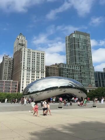 The media showcases a lively outdoor scene in Chicago, United States of America, centered around the iconic "Cloud Gate" sculpture, also known as "The Bean."

**Subjects:** The primary subjects are the people enjoying the public space and the large, reflective sculpture. Many individuals, including families with children, couples, and groups of friends, are scattered throughout the scene, walking, posing for pictures, and interacting with the environment. The sculpture itself is a dominant object, its polished, metallic surface reflecting the surrounding buildings, the sky, and the people. In the background, several tall, modern buildings form the urban skyline characteristic of a major city.

**Activity or Event:** The scene captures a moment of public enjoyment and tourism. People are actively engaged in exploring the park and interacting with the art installation. The atmosphere is relaxed and recreational, suggesting a popular tourist destination.

**Notable Details:** The time of day appears to be midday or early afternoon, indicated by the bright sunlight and clear blue sky with scattered white clouds. The weather seems pleasant and warm, suitable for outdoor activities. The architectural styles of the background buildings, ranging from older, ornate structures to sleek, contemporary skyscrapers, provide a strong sense of location in a bustling metropolitan area like Chicago. The reflections on "The Bean" offer dynamic visual information, mirroring the dynamic nature of the scene. The overall emotion conveyed is one of joy, wonder, and a sense of shared experience in a public urban space.

**Visible Text:** There is no visible text within the media file.