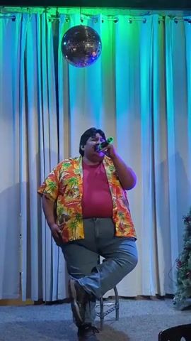 A heavy-set man is singing into a green toy microphone while sitting on a metal stool. He is wearing an orange Hawaiian shirt over a pink t-shirt and blue jeans, and brown work boots. A disco ball hangs from the ceiling. The backdrop is a light blue curtain. A Christmas tree is on the right.